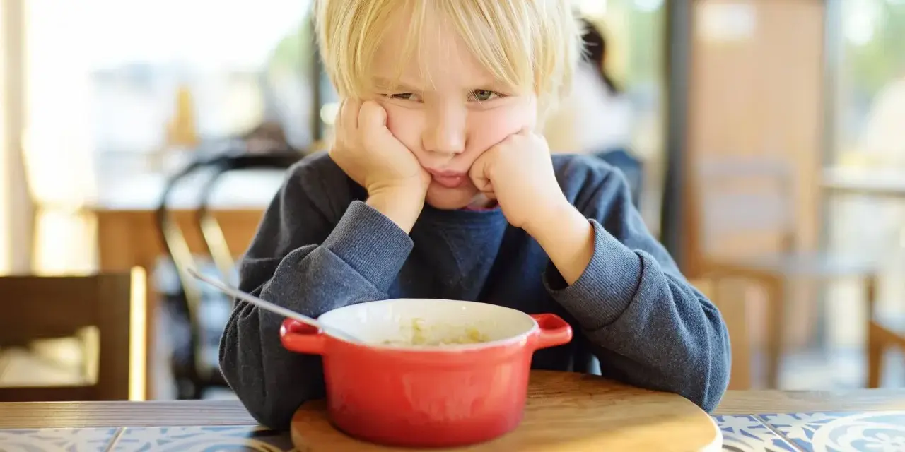 Ein Junge mit blonden Haaren stützt seinen Kopf auf die Hände und schaut lustlos auf eine Schüssel Porridge. Er isst nicht, obwohl er 10 Jahre alt ist.