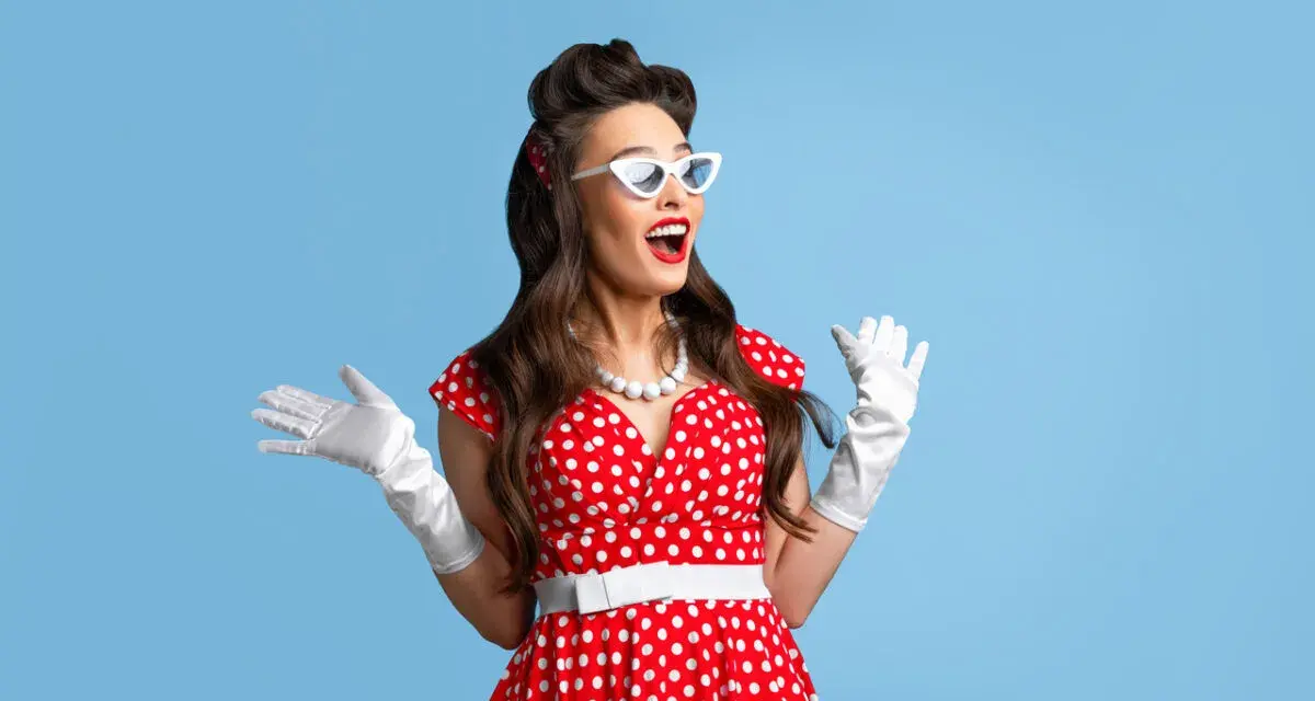 Kobieta w czerwonej sukience w grochy i białych rękawiczkach, z białymi okularami, prezentuje stylizacje pin up girl.