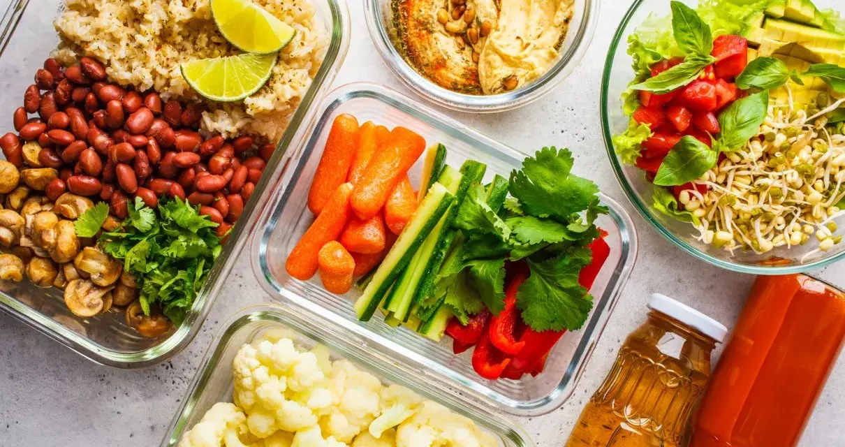 Gotowe pojemniki z jedzeniem na piknik: ryż z fasolą i pieczarkami, warzywa pokrojone w słupki, kalafior, sałatka z kiełkami i pomidorami, hummus i napoje.
