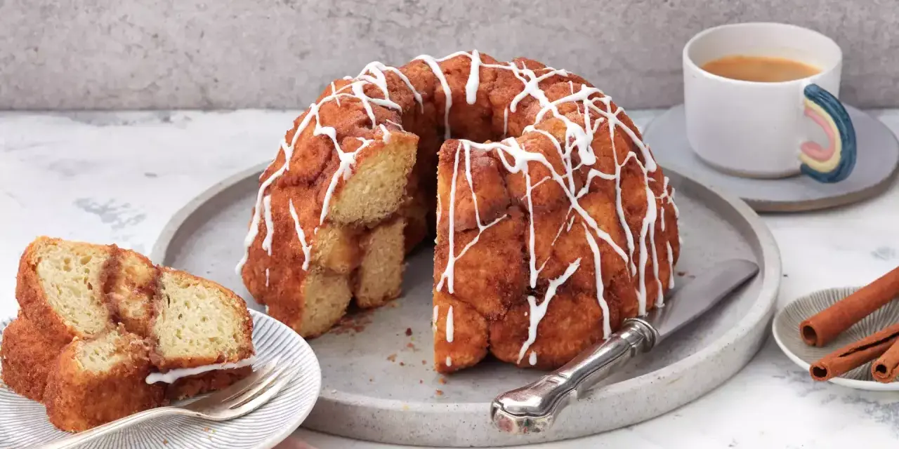 Ein köstlicher Monkey Bread mit Zimt und Zuckerguss, bereit zum Teilen. Daneben eine Tasse Kaffee und Zimtstangen.