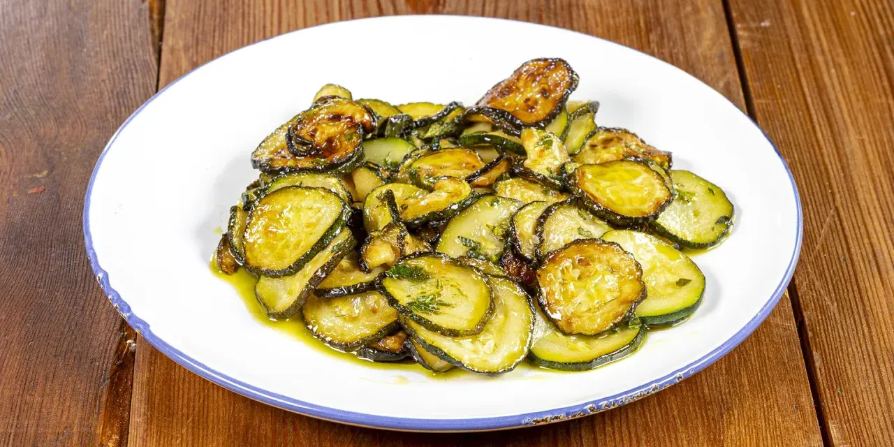 Zucchine alla poverella, fette sottili e dorate, condite con olio e erbe aromatiche, su un piatto bianco con bordo blu.