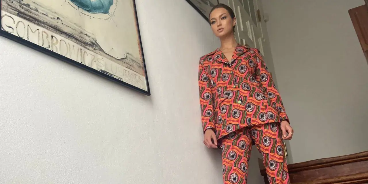Kobieta w kolorowym, wzorzystym garniturze, który przywodzi na myśl lata 70. stylizacje. W tle plakat teatralny.