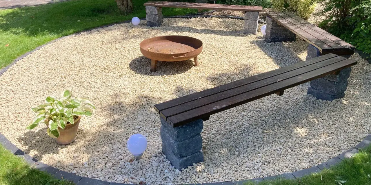Gemütliche Sitzecke im Garten mit Kiesboden, Feuerstelle und Holzbänken. Perfekt für laue Sommerabende.