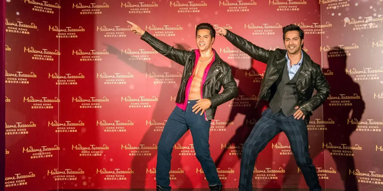 Dwie woskowe figury, jedna z odkrytym torsem, stoją na tle powtarzającego się logo Madame Tussauds w Hongkongu.