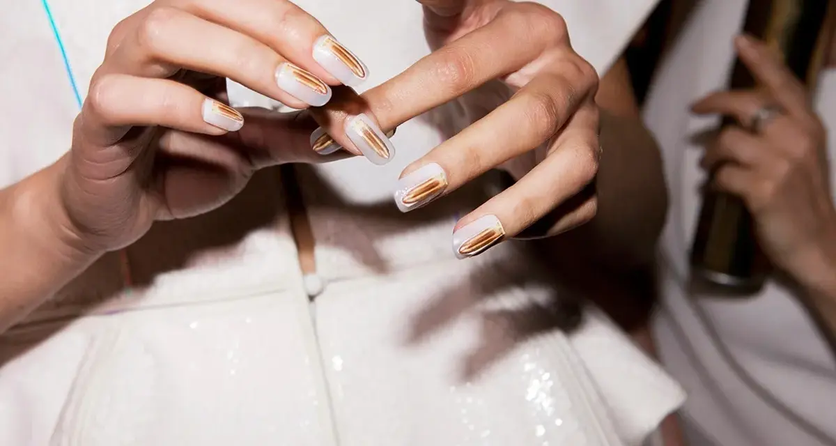 Eleganckie paznokcie przyklejane w białym kolorze ze złotymi zdobieniami. Dłonie prezentują nowoczesny manicure.