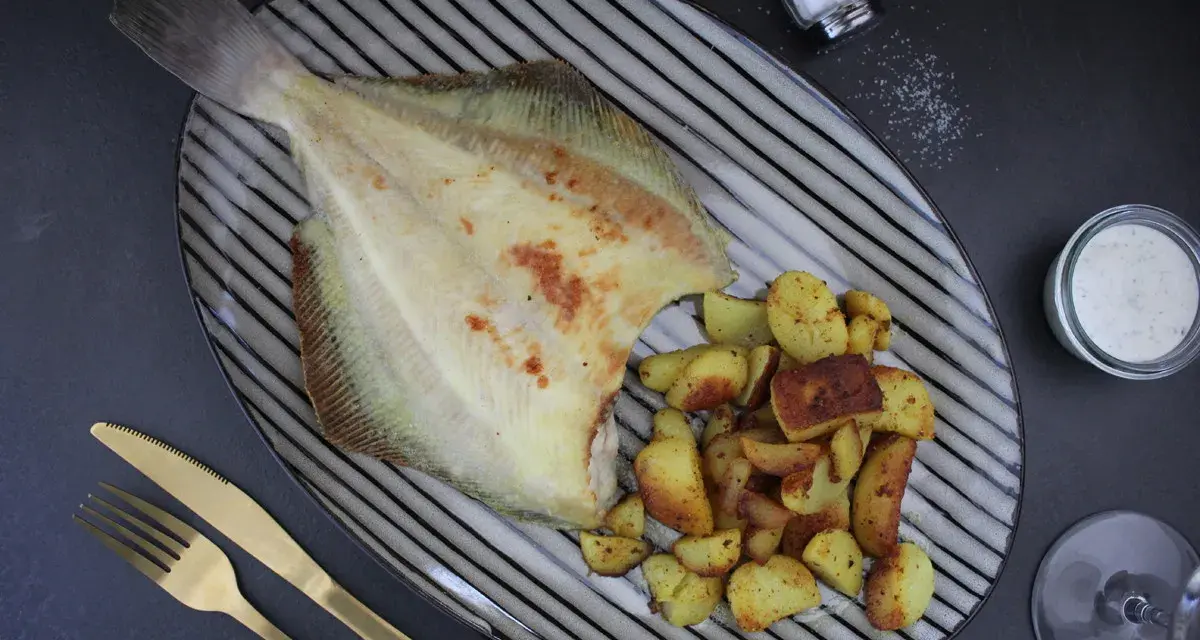 Gebratene Scholle Ostsee mit Bratkartoffeln und Dip auf einem gestreiften Teller, daneben ein Salzstreuer und goldenes Besteck.
