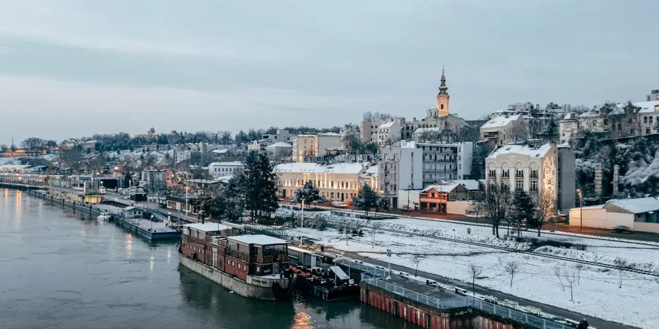 Widok na zimowe miasto nad rzeką z łodziami i ośnieżonymi budynkami.
