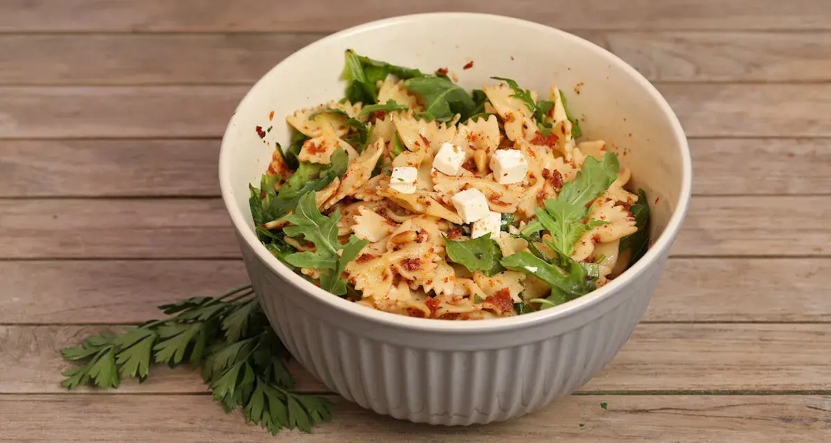 Schmackhafter Nudelsalat mit getrockneten Tomaten, Rucola und Feta in einer Schüssel auf Holzhintergrund.