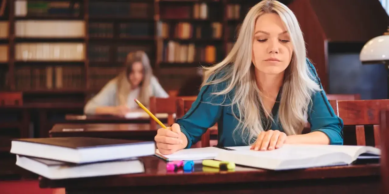 Kobieta z długimi blond włosami pisze w zeszycie, siedząc przy biurku w bibliotece.