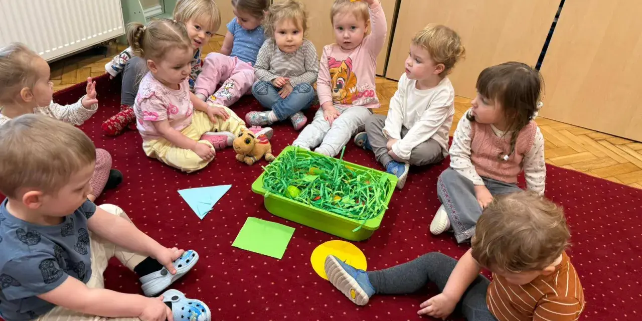 Dzieci siedzą na dywanie wokół zielonego pojemnika z zieloną trawą i jajkami.