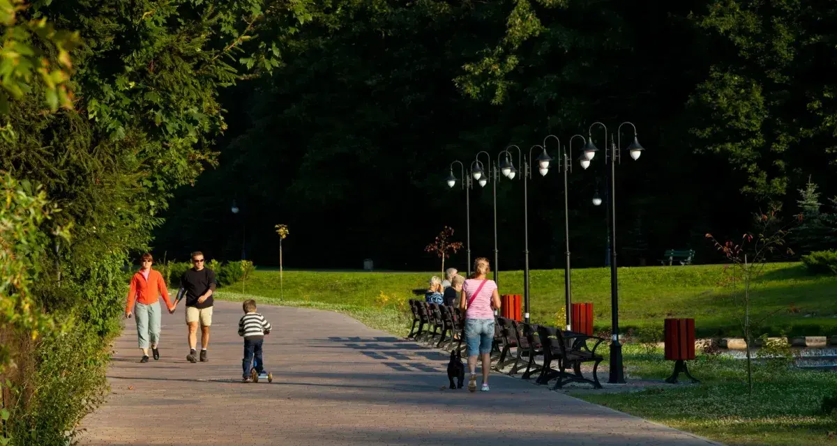 Rodzina spaceruje po parku z dzieckiem na hulajnodze i psem.