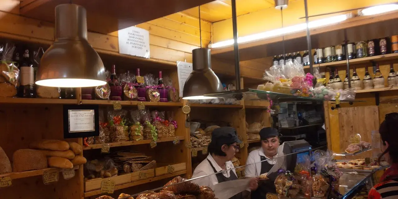 Interior of a bakery with shelves of bread, pastries, and bottles. Two women are behind the counter.