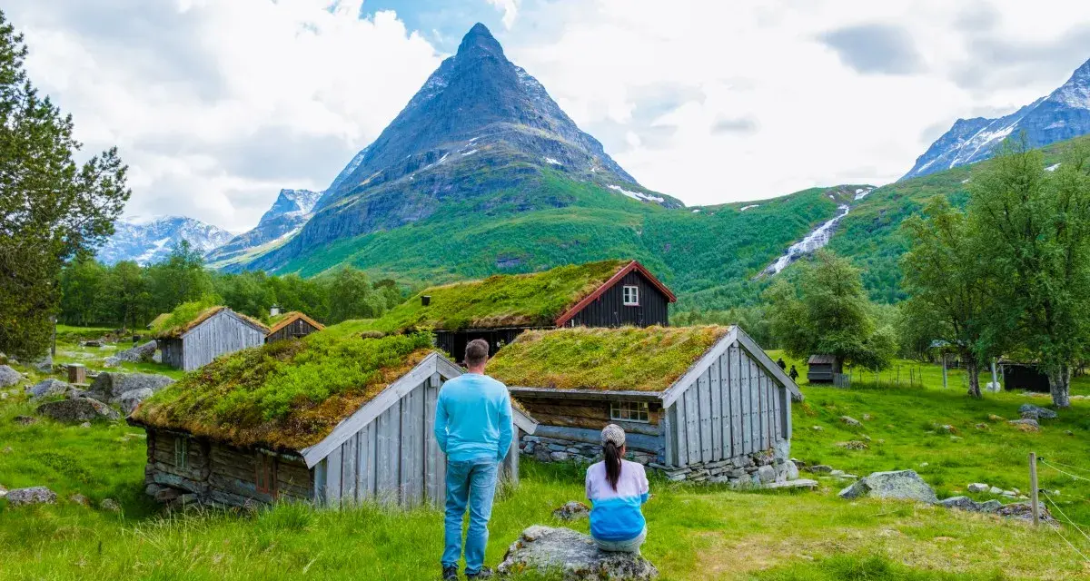 Para zwiedza ekuz w Norwegii, podziwiając tradycyjne chaty z zielonymi dachami i majestatyczne góry.