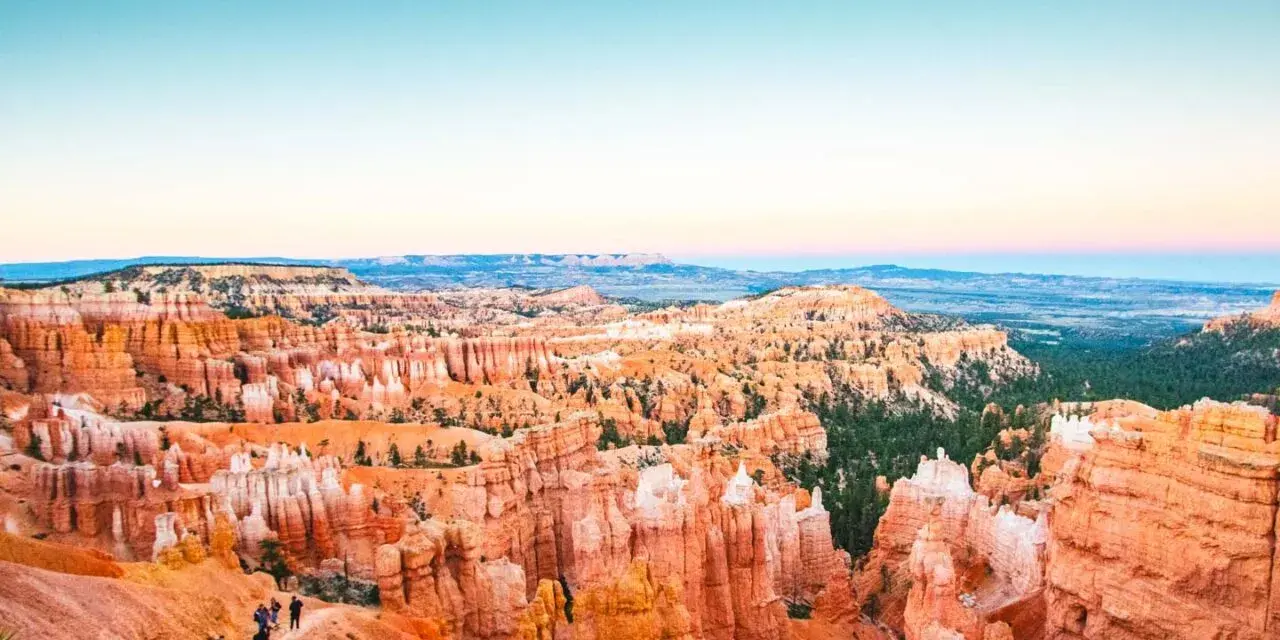 Zachód słońca nad Bryce National Park. Wąwóz pełen pomarańczowych hoodoos i zielonych drzew, z turystami na szlaku.