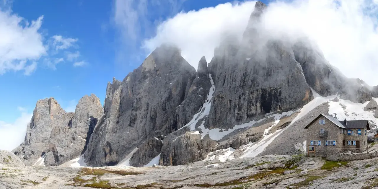 Rifugio in pietra ai piedi di imponenti vette rocciose, con neve residua e cielo azzurro. Un luogo incantevole al passo Valles.