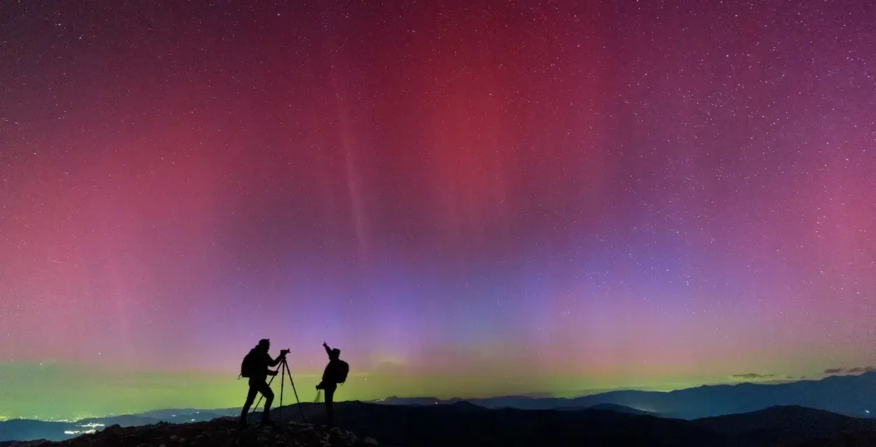 Due fotografi ammirano l'incredibile aurora boreale in Italia, con sfumature rosa e viola che danzano nel cielo stellato.