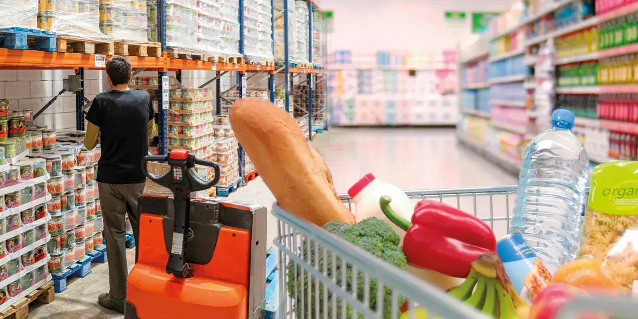 W magazynie FMCG, pracownik obsługuje wózek widłowy, podczas gdy wózek sklepowy pełen produktów czeka na zakupy.