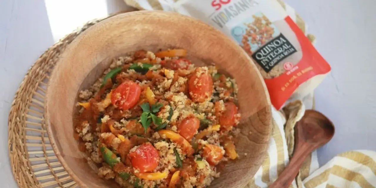Un tazón de madera lleno de quinoa con verduras coloridas: tomates cherry, pimientos verdes y amarillos. Cerca, un paquete de quinoa integral y roja.
