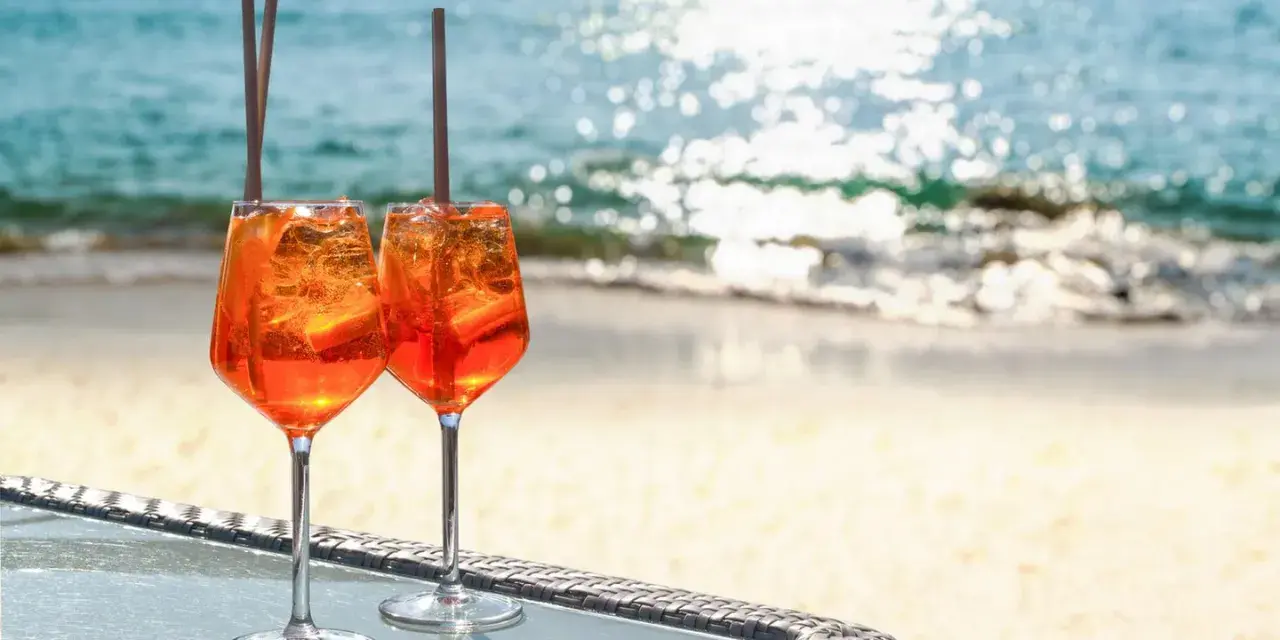 Zwei Aperol Spritz Gläser mit Eis und Orangenscheiben stehen am Strand. So trinkt man Aperol Spritz in Italien!