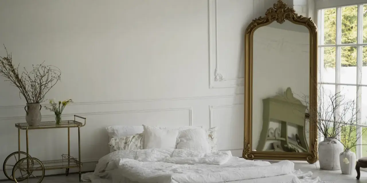 Elegancka sypialnia z łóżkiem w bieli, ozdobiona sztukaterią przypominającą lamperię angielską. Złote lustro i wózek dodają blasku.