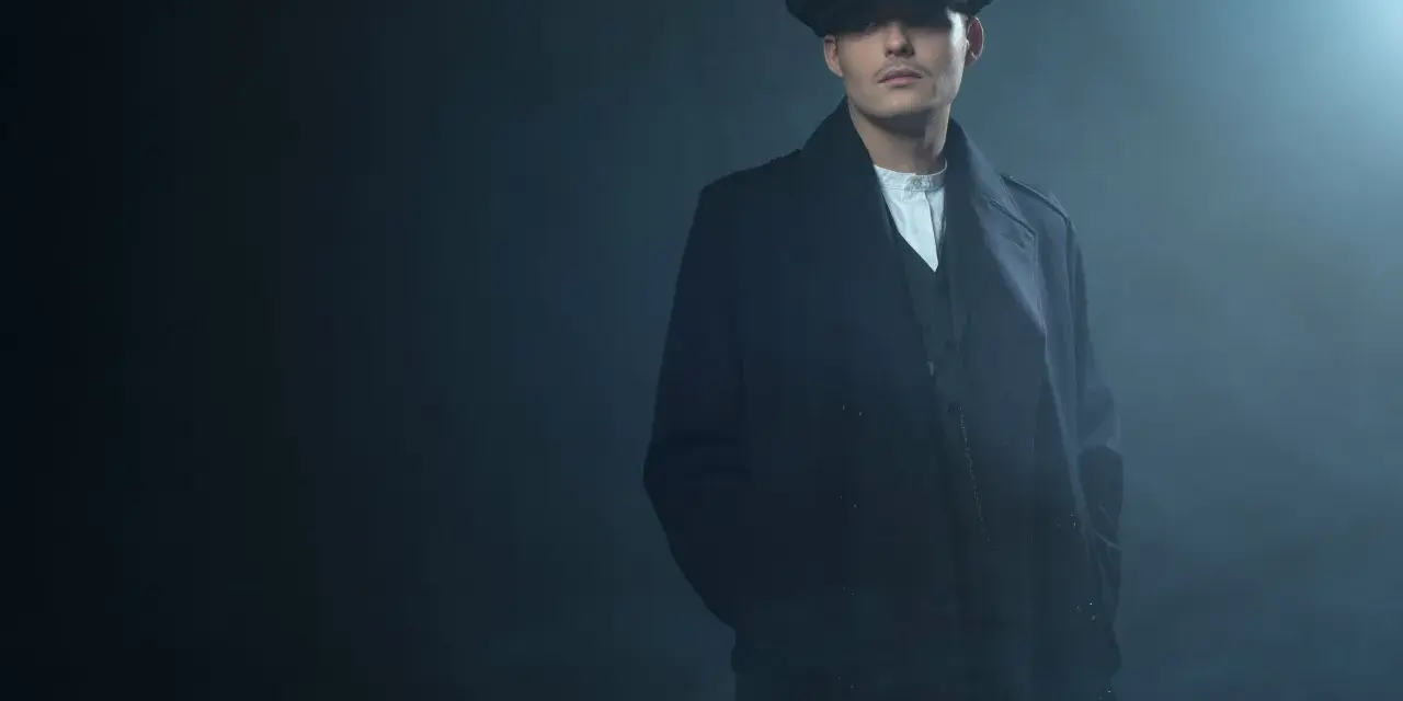 Mężczyzna w kaszkiecie i płaszczu, z charakterystycznym wąsem, emanuje chłodnym spojrzeniem w stylu Peaky Blinders.
