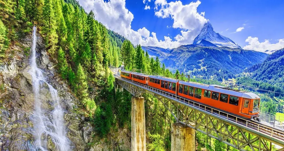 An orange train crosses a bridge over a waterfall, on its way from Zurich to Andermatt. Lush green trees and mountains surround the scenic route.