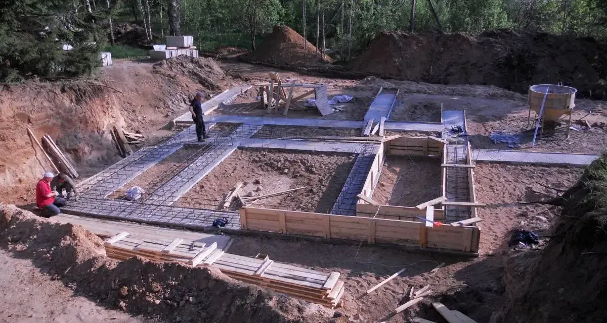 Budowa fundamentów: zbrojenie i szalunki czekają na chudy beton. W tle las i plac budowy.