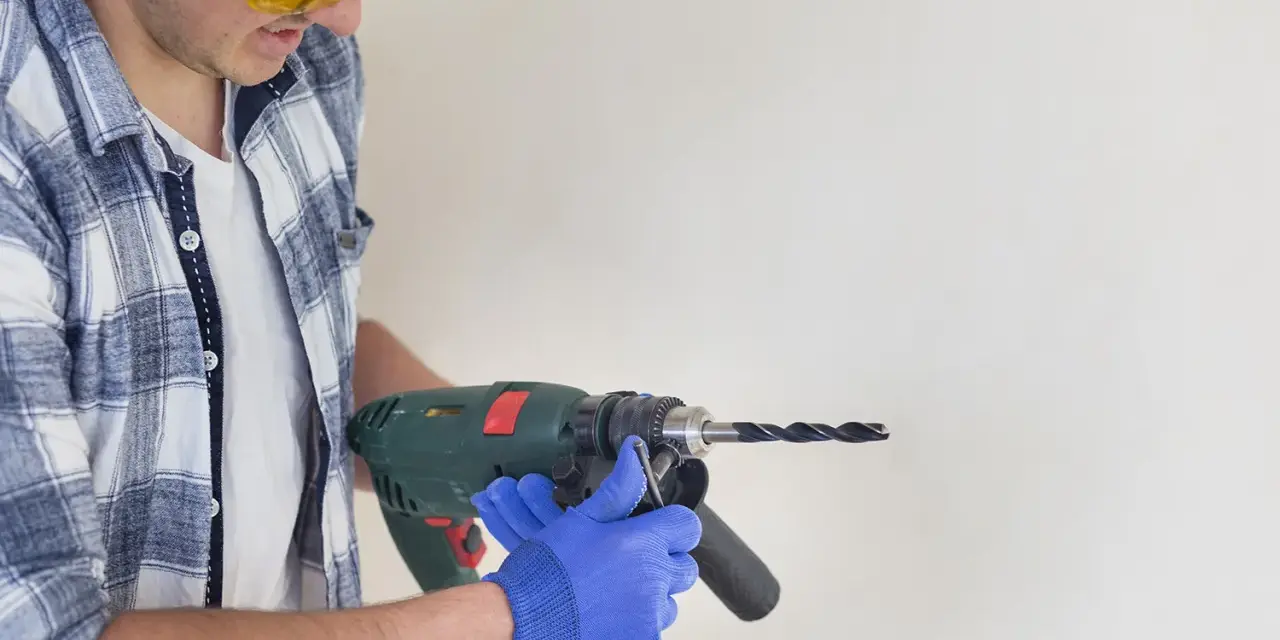 Man in safety glasses and blue gloves holding a drill to a white surface.