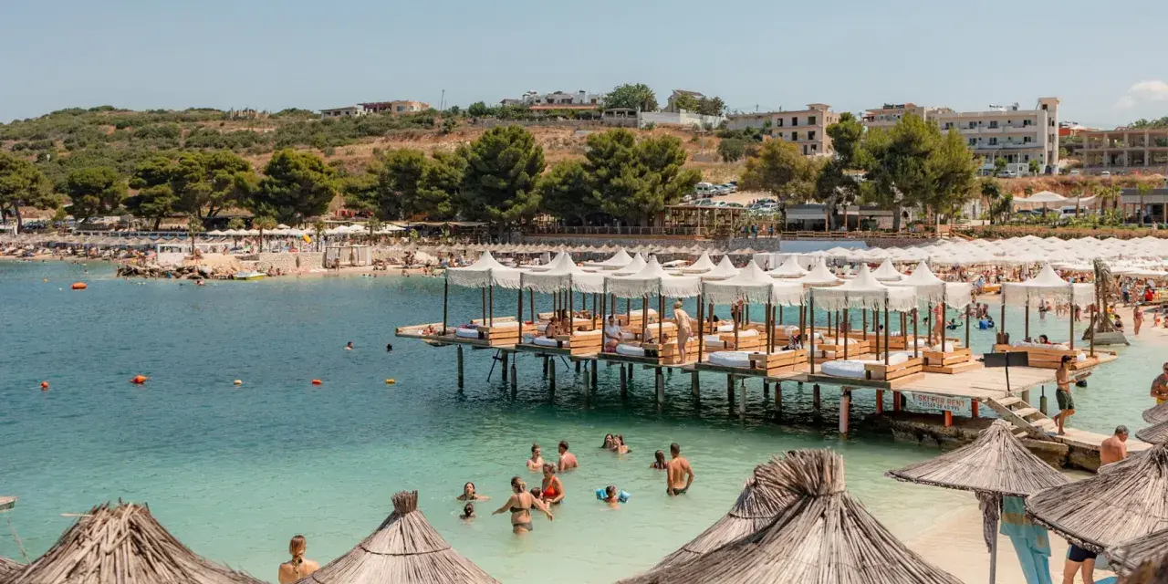 Słoneczny dzień na Ksamil Beach. Ludzie kąpią się w turkusowej wodzie, relaksują się na leżakach pod białymi parasolami i na drewnianych pomostach.