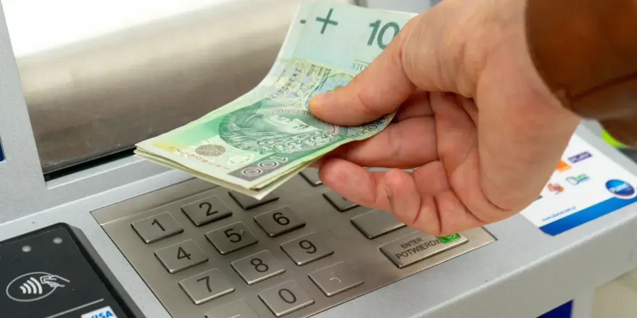Dłoń wkłada banknoty 100 zł do bankomatu. Zastanawiasz się, ile można wpłacić na konto?