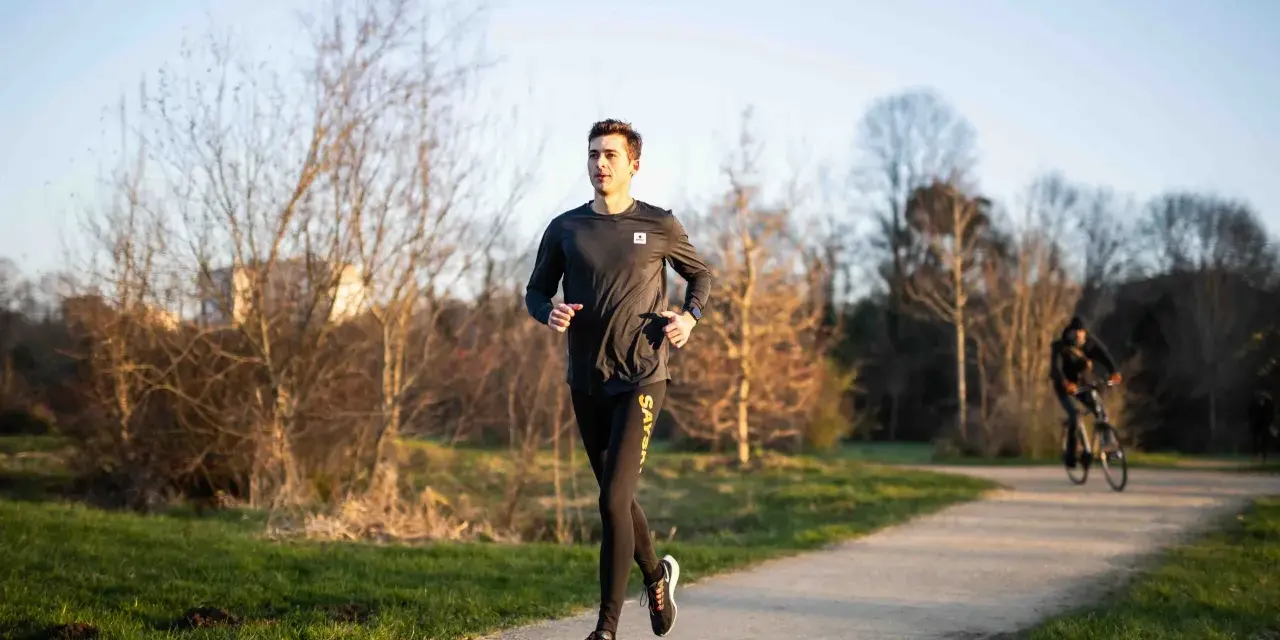 Un jeune homme s'entraîne à la course à pied sur un chemin ensoleillé, entouré d'arbres dénudés. Un cycliste passe en arrière-plan.