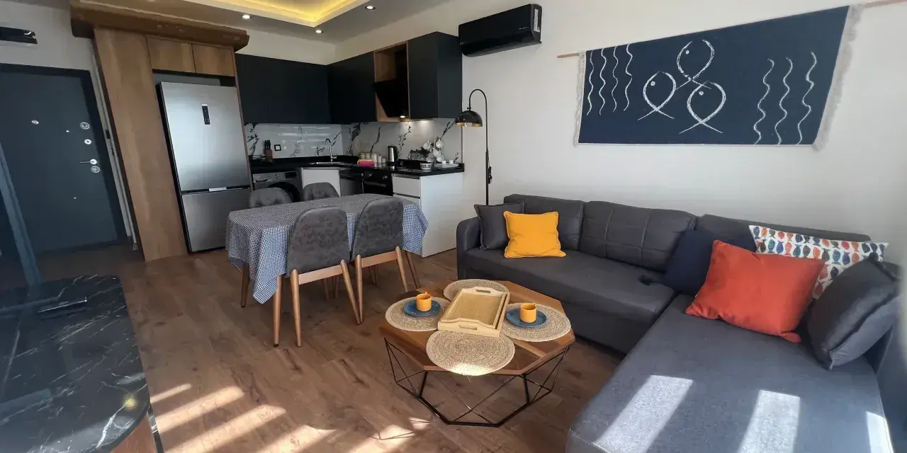 Nowoczesny salon z aneksem kuchennym. Wygodna kanapa, stolik kawowy, stół z krzesłami. W tle lodówka i sprzęty kuchenne. Idealne miejsce na relaks, z myślą o komforcie gości.