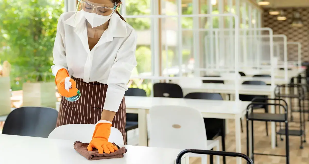 Employée portant masque et visière nettoie une table, respectant la réglementation hygiène cantine scolaire. Des séparateurs sont installés.