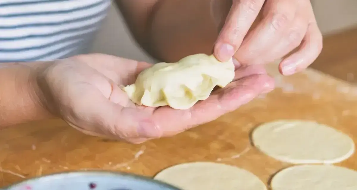 Ręce formują pierogi ruskie na drewnianej desce. W tle miska z wiśniami.