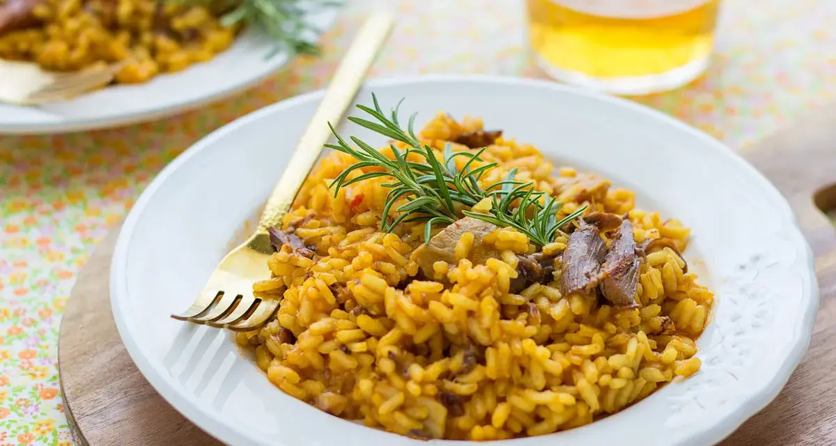Delicioso arroz con setas y carne, servido en plato blanco con tenedor dorado y ramita de romero.