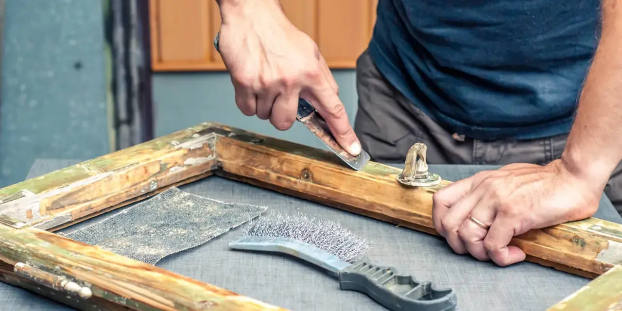 Uomo usa raschietto per rimuovere vecchia vernice da telaio in legno, mostrando come sverniciare il legno con attrezzi.