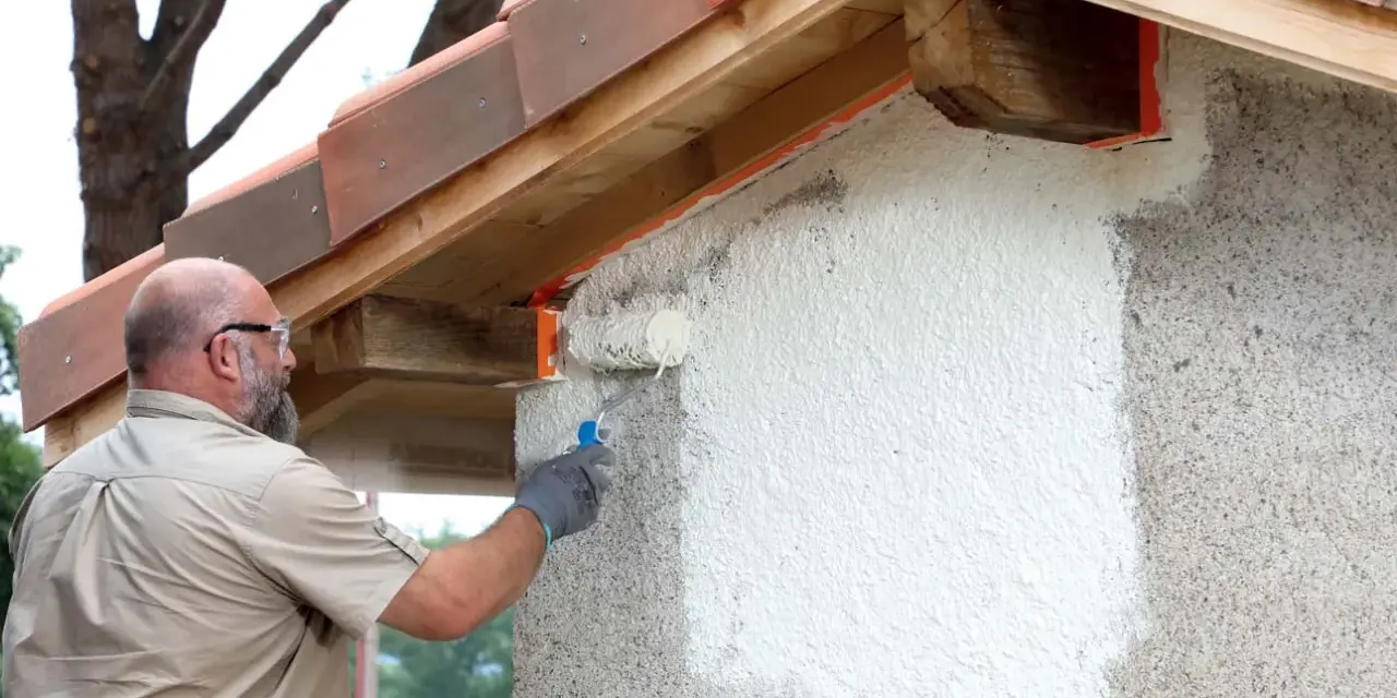 Un homme applique une peinture sur mur béton extérieur avec un rouleau. La rénovation progresse sous le toit de tuiles.