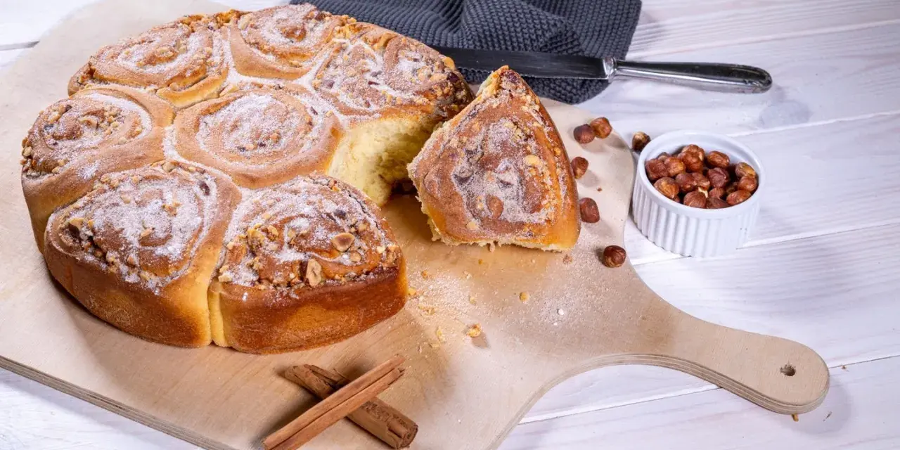 Ein köstliches Zimtschnecken Rezept: goldbraune, mit Nüssen und Puderzucker bestreute Schnecken auf einem Holzbrett, daneben Zimtstangen und Haselnüsse.