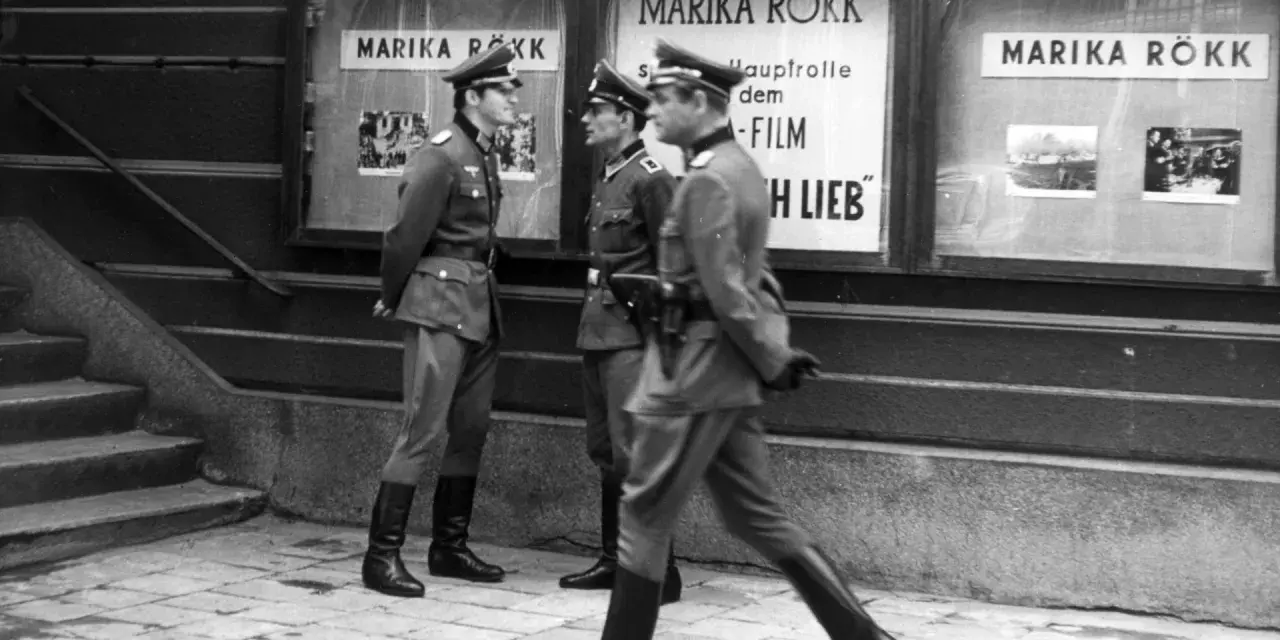 Trzej żołnierze w mundurach stoją przed kinem z plakatem Mariki Rökk. Stawka większa niż życie, teatr historii.