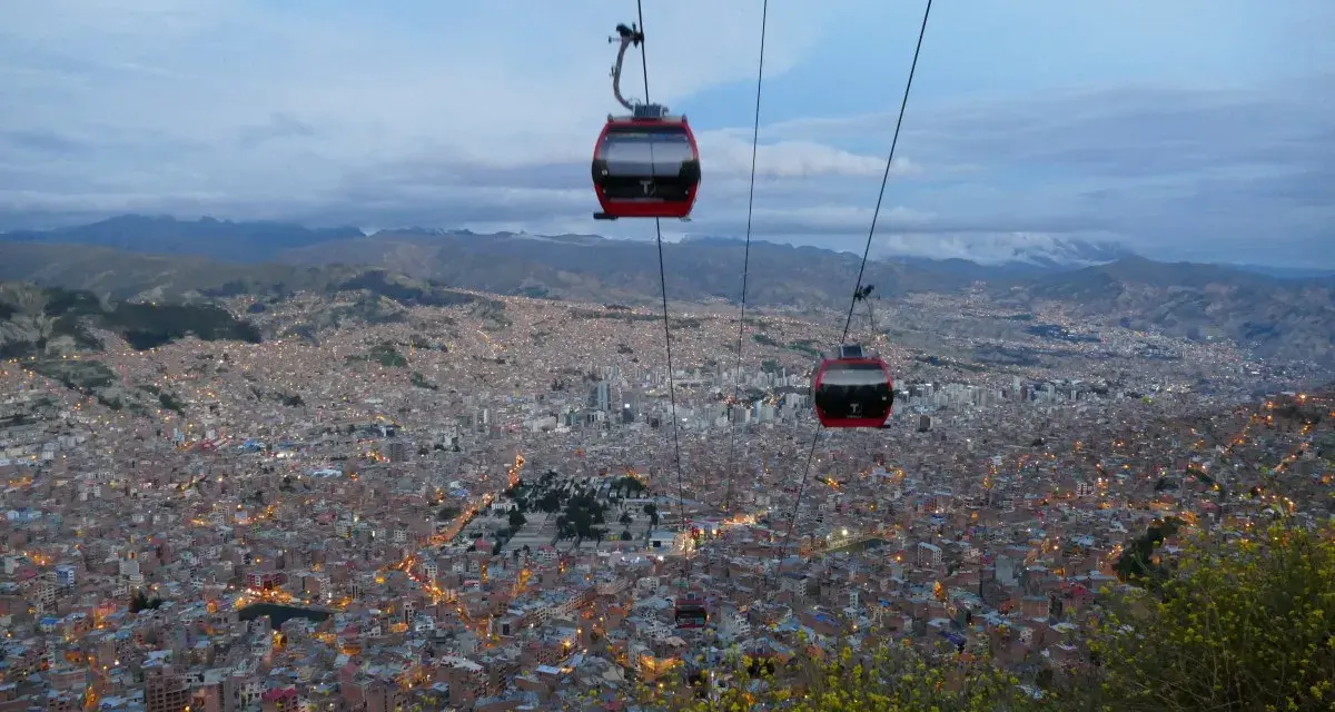 Red la paz gondola cabins glide over a sprawling city at dusk, with lights twinkling below and mountains in the distance.
