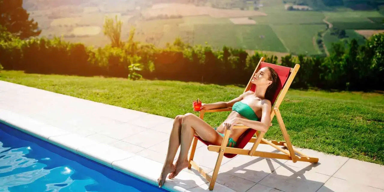 Donna rilassata su una sdraio, con drink, vicino a una piscina. Il sole splende, ideale per chi vuole sapere come abbronzarsi velocemente.