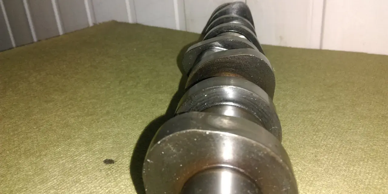 Metalowy, wytarty wałek rozrządu leży na zielonym materiale. Widać ślady zużycia na jego krzywkach.