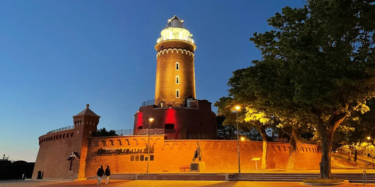 Wieczorny spacer przy latarni morskiej Kołobrzeg. Oświetlona ceglana budowla z czerwonymi akcentami i drzewa tworzą magiczny klimat.