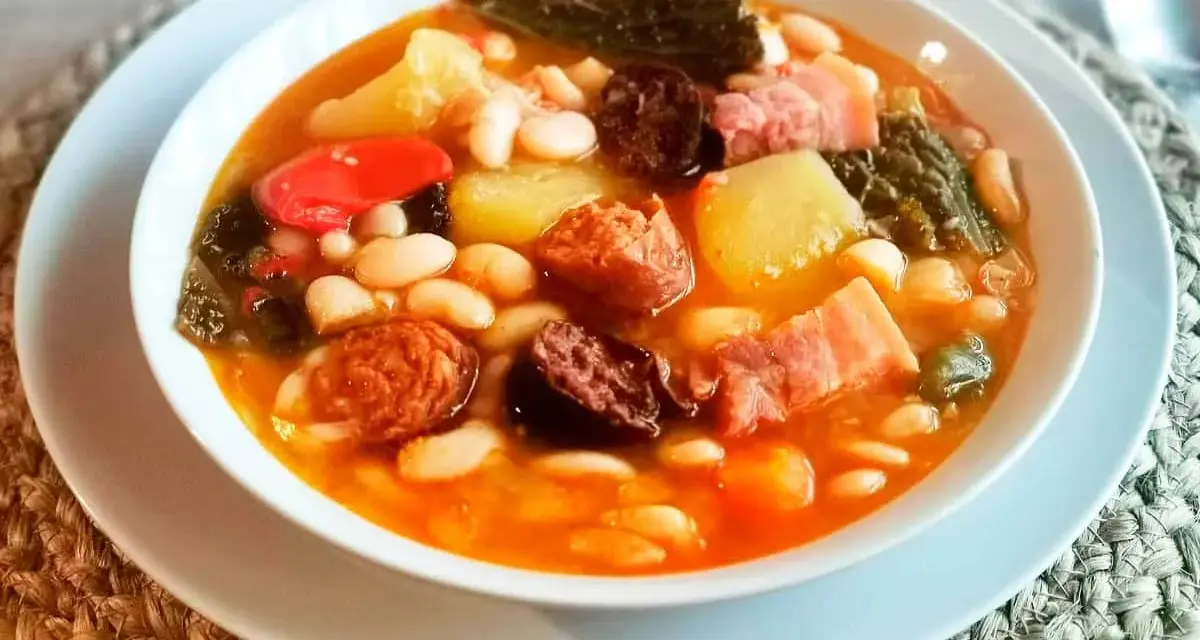 Un reconfortante cocido montañés, rebosante de alubias blancas, patatas, chorizo, morcilla y tocino, servido en un cuenco blanco.