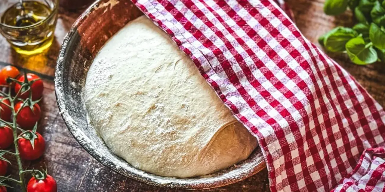 Frischer Pizzateig ruht in einer Schüssel, bedeckt mit einem rot-weiß karierten Tuch. Zutaten für ein Pizzateig Rezept wie Tomaten und Öl liegen bereit.
