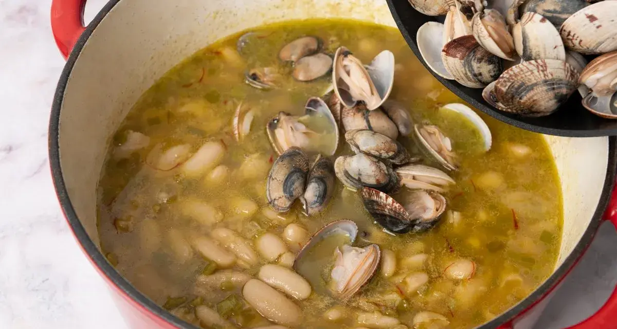 Alubias cremosas con almejas en una olla roja, añadiendo más almejas de una sartén.