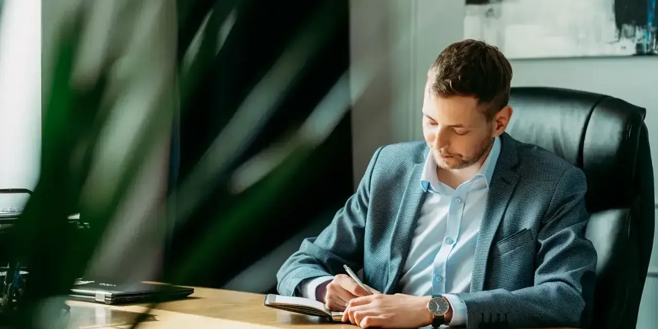 Mężczyzna w garniturze siedzi przy biurku i pisze w notatniku.
