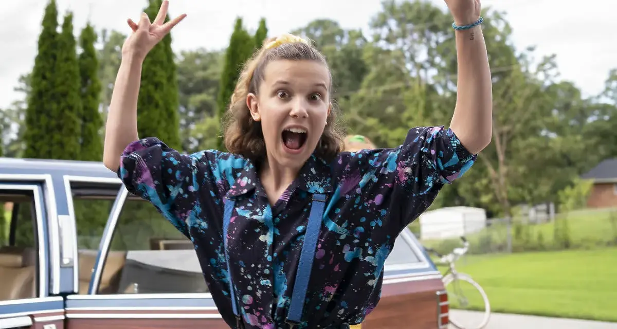 Millie Bobby Brown w kolorowej koszuli i szelkach, radośnie pozuje przy samochodzie. Jej filmy, seriale i programy to gwarancja emocji!