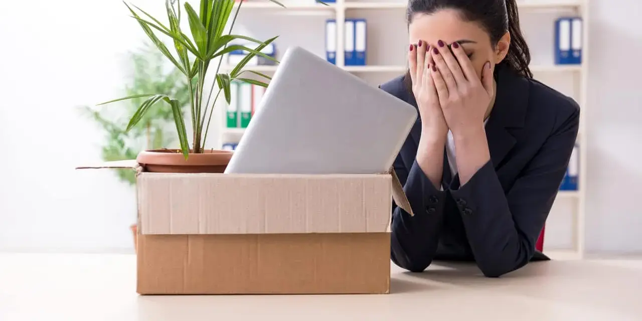 Verzweifelte Frau mit Karton und Laptop im Büro. Sie muss eine innerbetriebliche Versetzung ablehnen und ist sichtlich mitgenommen.