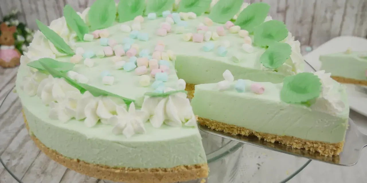 Eine helle, cremige Philadelphia Torte Waldmeister mit bunten Marshmallows und grünen Blättern verziert. Ein Stück wird gerade herausgeschnitten.